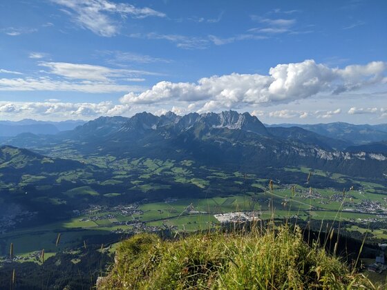 Aussicht auf das Kaisergebirge Blick von einem grasbewachsenen Gipfel auf ein weites Tal und ein schroffes Gebirgsmassiv unter blauem Himmel.