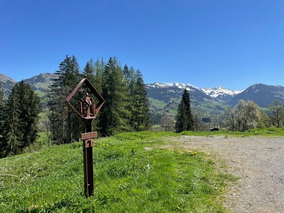 Station am Kreuzweg Ein hölzernes Marterl auf einer grünen Wiese vor einem Bergpanorama mit schneebedeckten Gipfeln.