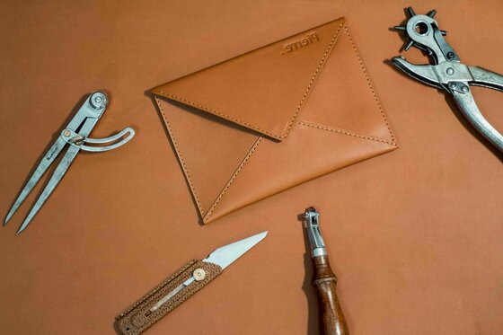 Lederhandwerk My Pierre Braune Ledertasche in Briefumschlagform umgeben von Lederwerkzeugen wie Lochzange und Zirkel auf braunem Grund.