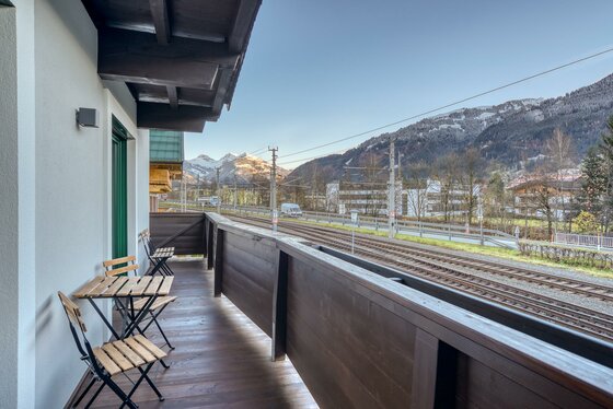 Holzbalkon mit Sitzgruppe und Blick auf Bahngleise sowie schneebedeckte Berge.