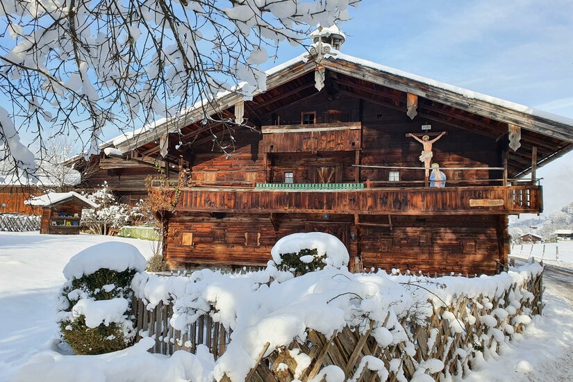 Verschneites historisches Bauernhaus aus dunklem Holz mit Kruzifix an der Fassade und schneebedecktem Zaun davor.