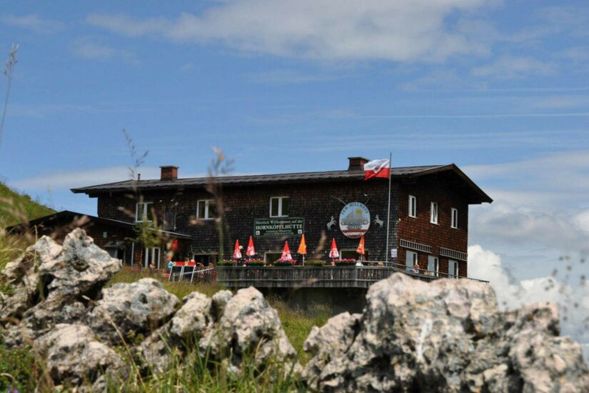 Zweistöckige Berghütte aus dunklem Holz mit Sonnenschirmen auf der Terrasse hinter Felsen.