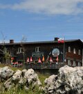 Zweistöckige Berghütte aus dunklem Holz mit Sonnenschirmen auf der Terrasse hinter Felsen.