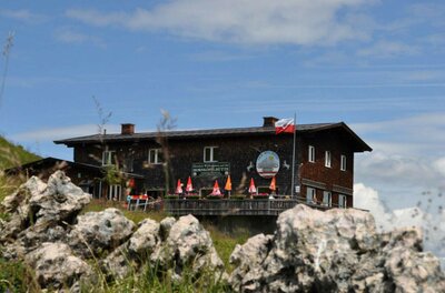 Zweistöckige Berghütte aus dunklem Holz mit Sonnenschirmen auf der Terrasse hinter Felsen.