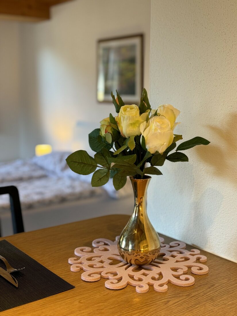 Weiße Rosen in einer goldenen Vase auf einem Holztisch vor einem unscharfen Schlafbereich.