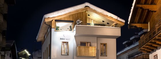 Beleuchtetes alpines Gebäude bei Nacht mit schneebedecktem Dach und Holzfassade im Giebelbereich.