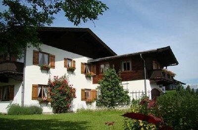 Zweistöckiges alpines Wohnhaus mit weißen Wänden, Holzfensterläden und Balkonen vor einer Wiese.