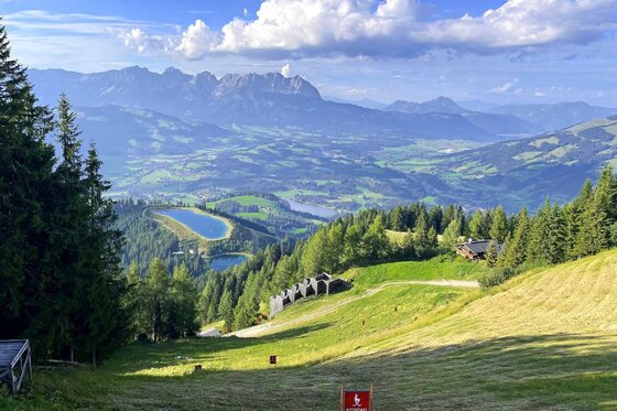 Alpines Panorama mit Speichersee Blick von einer Almwiese auf ein Tal mit Speichersee und Gebirgskette im Hintergrund.
