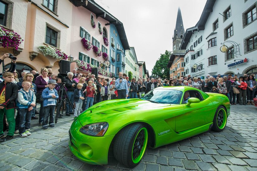 Ein neongrüner Sportwagen fährt auf Kopfsteinpflaster an einer Menschenmenge in einer historischen Altstadt vorbei.