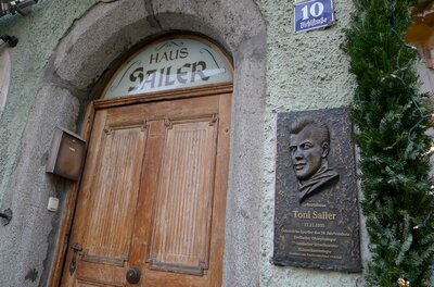 Eine alte Holztür mit dem Schild "Haus Sailer" und einer Gedenktafel für Toni Sailer. Die Wand ist in einem sanften Grün gestrichen.