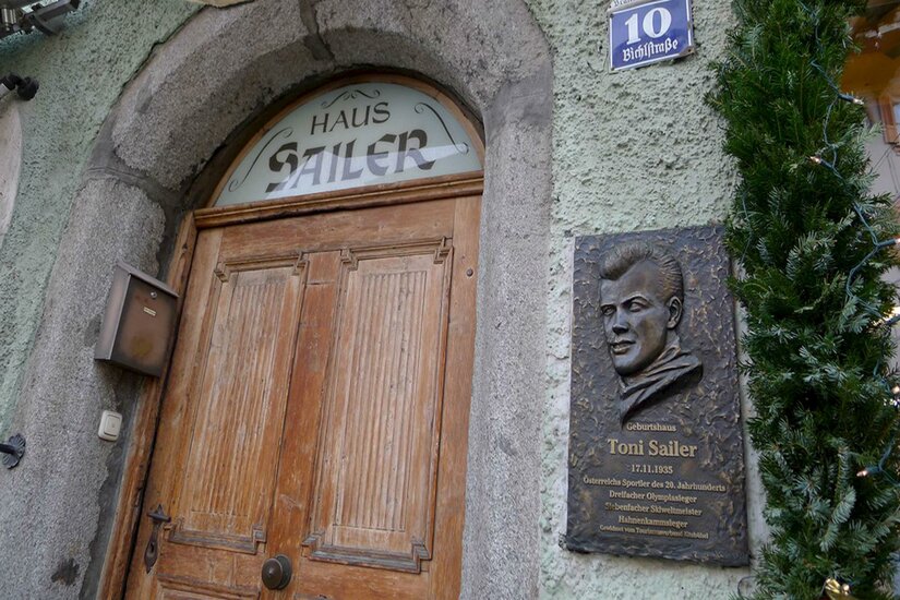Eine alte Holztür mit dem Schild "Haus Sailer" und einer Gedenktafel für Toni Sailer. Die Wand ist in einem sanften Grün gestrichen.