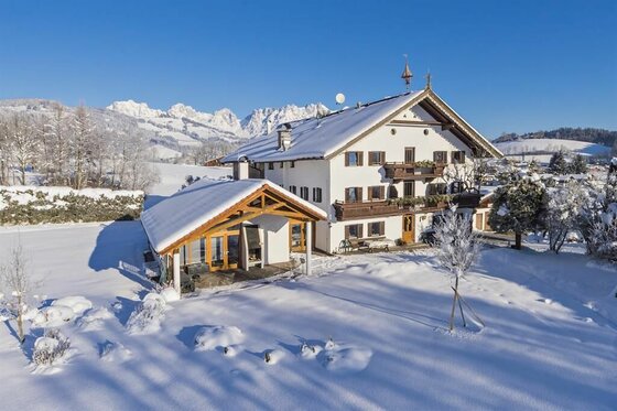 Pension Thainerhof im Winter Verschneites, mehrstöckiges Alpenhaus mit Holzbalkonen vor schneebedeckten Bergen unter blauem Himmel.