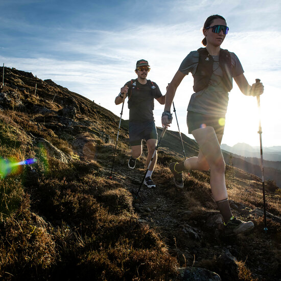In der goldenen Abendsonne rennen zwei Personen mit Wanderstöcken über einen malerischen Bergpfad, umgeben von sanften Hügeln.