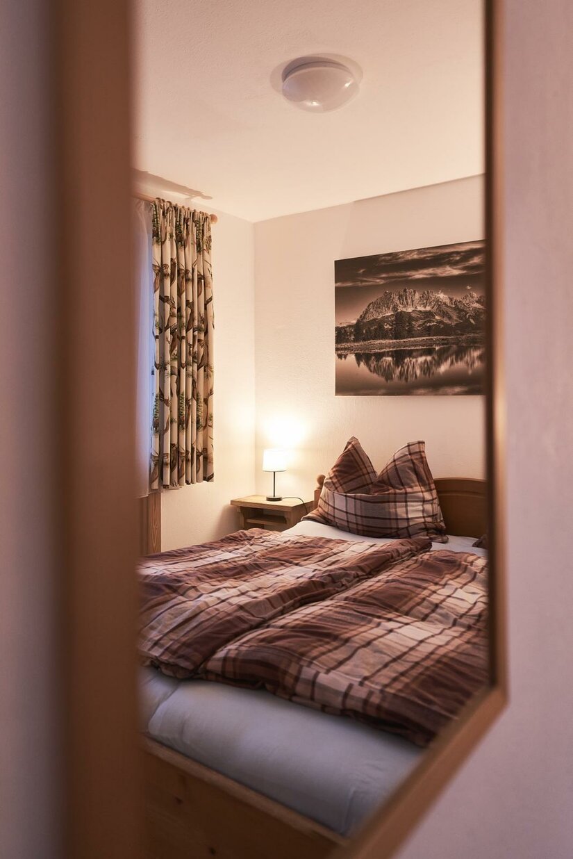Schlafzimmer Ferienwohnung Hahnenkammblick Blick durch einen Türrahmen auf ein Doppelbett mit karierter Bettwäsche und ein Bergbild an der Wand.