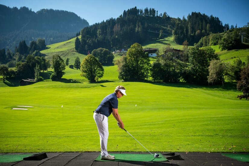 Ein Golfer steht auf einer Driving Range inmitten einer grünen Landschaft. Im Hintergrund sind sanfte Hügel und Bäume zu sehen.