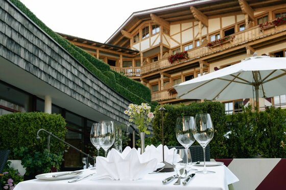 Terrasse Hotel Kaiserhof Kitzbühel Gedeckter Tisch auf einer Hotelterrasse mit Weingläsern und Blick auf ein Holzgebäude im alpinen Stil.