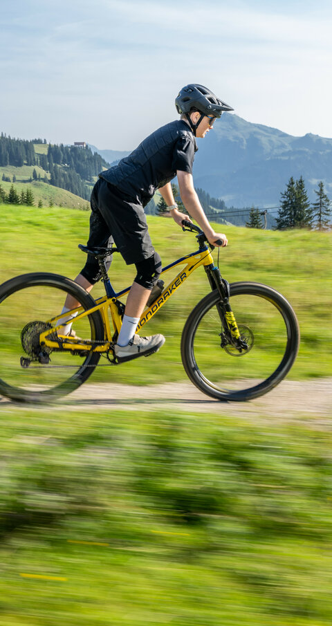 Ein junger Mountainbiker fährt im MTB Family Area Streiteck bei den 360 Flow Trails in Kitzbühel mit einem gelben Mountainbike über einen schmalen Weg in der grünen Berglandschaft.