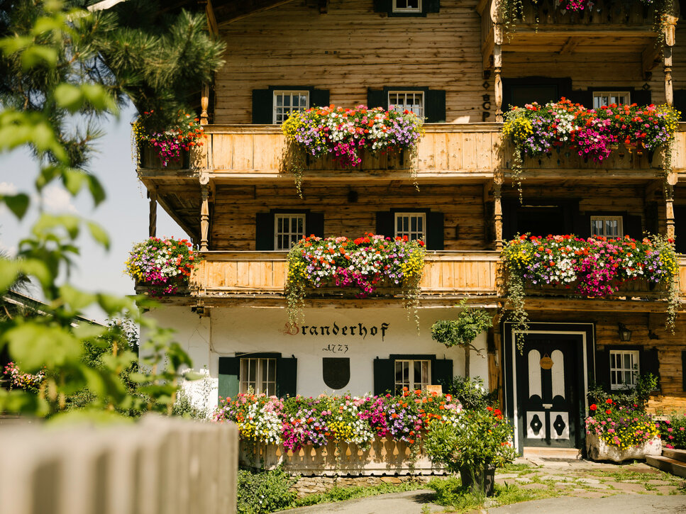 Blick auf ein rustikales Bauernhaus mit üppigen Blumenarrangements an den Balkonen, umgeben von grüner Vegetation.