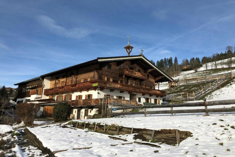 Henntalhof im Winter Großes Holzhaus im alpenländischen Stil mit Balkonen auf einer verschneiten Wiese unter blauem Himmel.