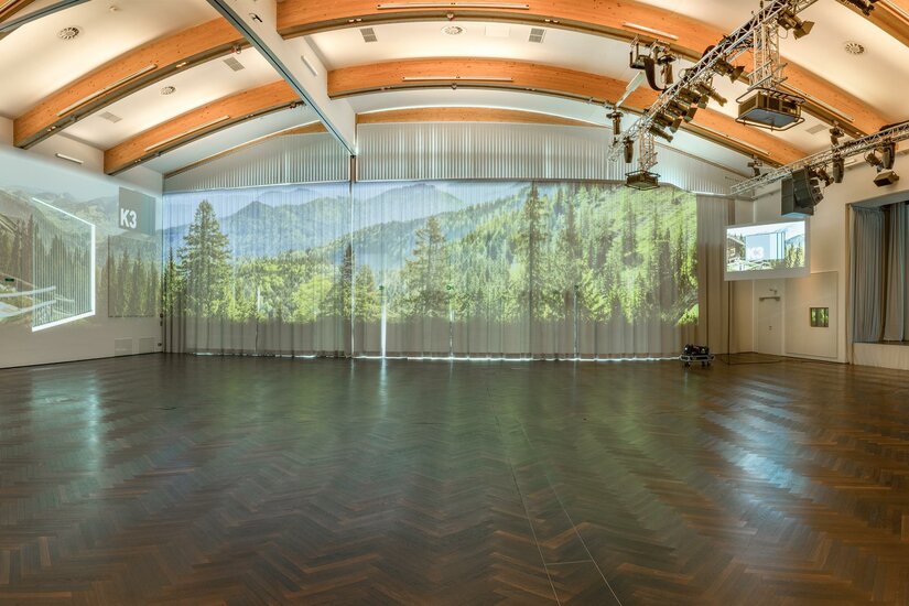 K3 - KitzKongress Ein moderner Veranstaltungsraum mit Holzdecke und großen Fenstern. Auf den Wänden sind Landschaftsprognosen und Präsentationen projiziert.