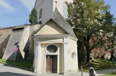 Ein kleines, historisches Gebäude mit einem spitzen Dach steht neben einer Kirche. Die Umgebung ist grün, mit Bäumen und einem ruhigen Weg.