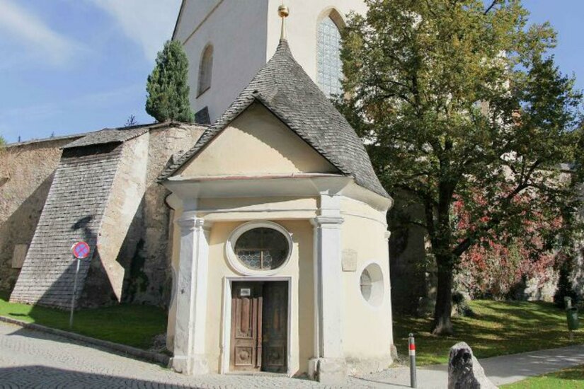 Johann Nepomuk Kapelle Ein kleines, historisches Gebäude mit einem spitzen Dach steht neben einer Kirche. Die Umgebung ist grün, mit Bäumen und einem ruhigen Weg.