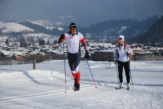 Zwei Personen beim Skilanglauf auf einer präparierten Loipe vor einer verschneiten Ortschaft.
