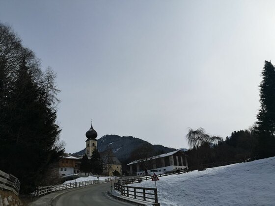 Blick auf eine asphaltierte Straße, eine Kirche mit Zwiebelturm und schneebedeckte Hänge vor bewaldeten Bergen.