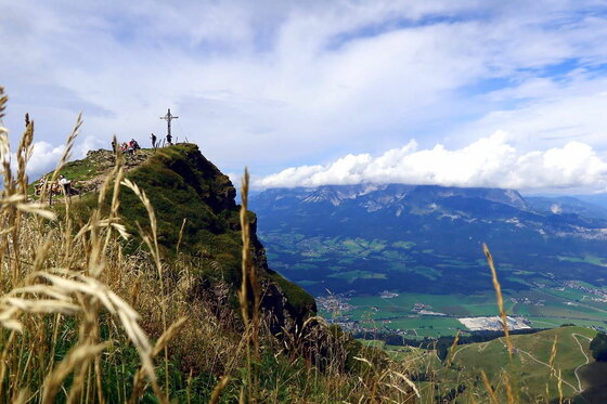 Gipfelkreuz auf einem grasbewachsenen Bergkamm mit Wanderern und Blick ins Tal.