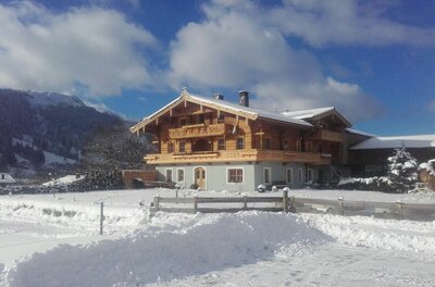 Ein traditionelles Holzhaus mit Balkonen in einer verschneiten Winterlandschaft vor Bergen.