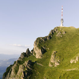 Ein malerischer Berg mit einem Sendemast, der über die sanften Hügel und das grüne Gras auf dem Hang ragt, unter einem blauen Himmel.