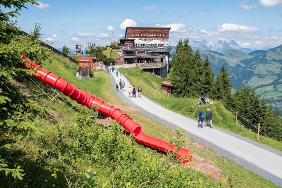 Rote Röhrenrutsche neben einem asphaltierten Wanderweg an einem Berghang mit Berggasthof im Hintergrund.