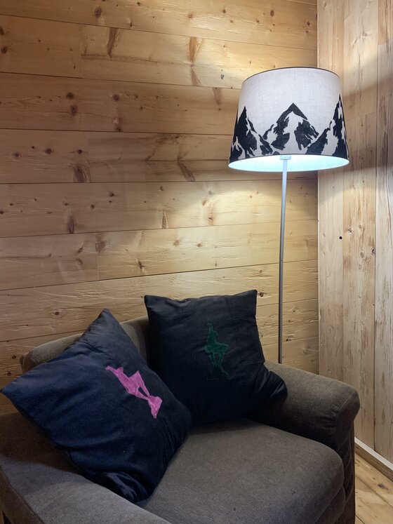 Kleiner Loungebereich Sessel mit zwei dunklen Kissen vor einer Holzwand neben einer Stehlampe mit Bergmotiv.