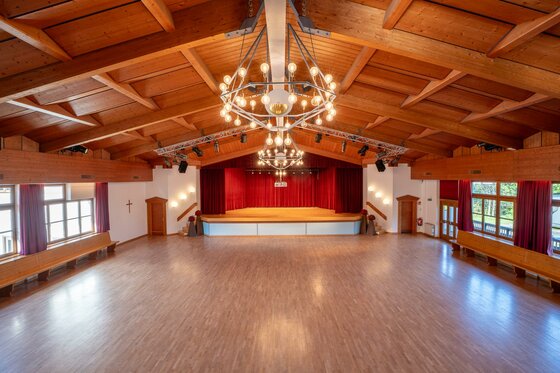 Saal <p>Ein großer Veranstaltungsraum vom Kulturhaus Reith mit einer Holzdecke und einem Bühnenbereich. Der Raum ist gut beleuchtet und hat rote Vorhänge.</p>