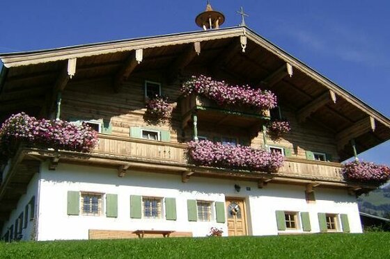 Traditionelles Alpenhaus mit weißer Fassade, Holzobergeschoss und blumengeschmückten Balkonen vor blauem Himmel.