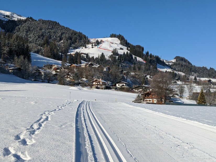 Präparierte Langlaufloipe im Schnee vor einer Siedlung und bewaldeten Bergen unter blauem Himmel.