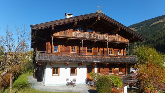 Mehrstöckiges Holzhaus mit Balkonen und weißem Erdgeschoss vor bewaldetem Berg.