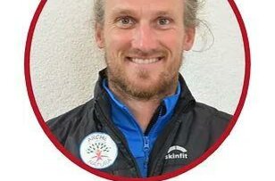 Bergwanderführer Ein lächelnder Mann mit langen, blonden Haaren trägt eine schwarze Jacke und ein blaues Underlayer. Der Hintergrund ist hell und der Mann ist in einem roten Kreisrahmen eingefasst.