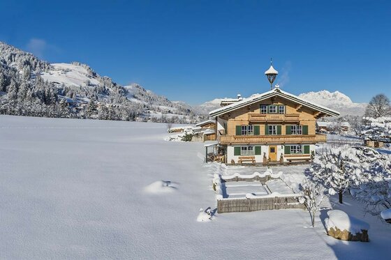 Traditionelles Holzhaus in einer verschneiten Alpenlandschaft unter blauem Himmel.