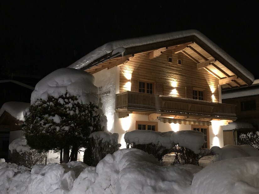 Haus Hagleitner im Winter Beleuchtetes Holzhaus bei Nacht mit schneebedecktem Dach und tief verschneiten Hecken im Vordergrund.