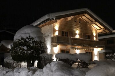 Beleuchtetes Holzhaus bei Nacht mit schneebedecktem Dach und tief verschneiten Hecken im Vordergrund.