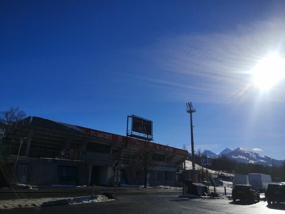 Außenansicht eines Stadions mit Flutlichtmast vor schneebedeckten Bergen unter blauem Himmel.
