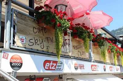 Balkon mit Holzverkleidung, roten Geranien, Sonnenschirmen und Werbeschildern von Sportgeschäften.