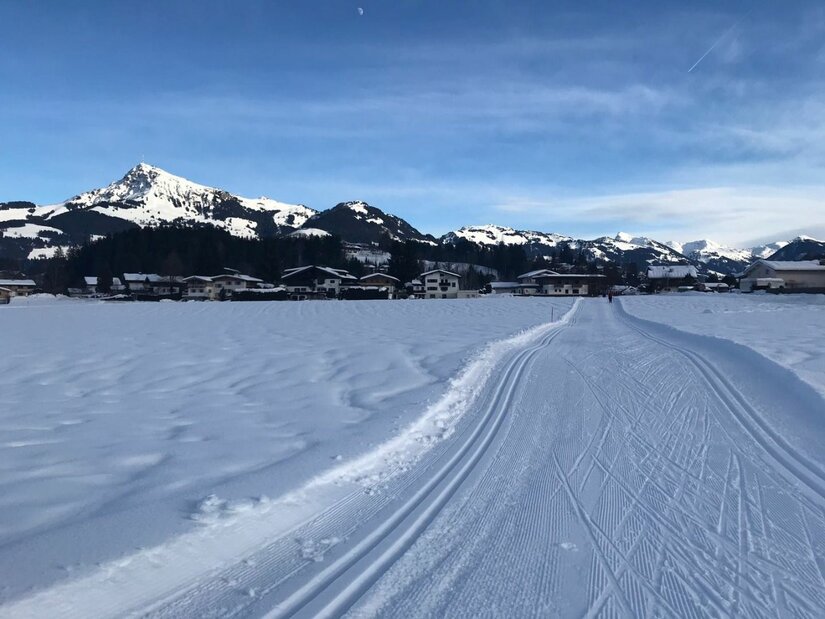 Präparierte Langlaufloipe auf einer verschneiten Ebene vor einer Bergkulisse und Häusern.