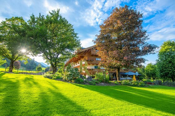 Außenansicht Rasmushof Hotel Kitzbühel Mehrstöckiges Holzhotel mit Balkonen hinter einer grünen Wiese und großen Laubbäumen bei Sonnenschein.