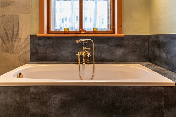 Weiße Badewanne mit goldener Armatur vor einem Fenster mit dunkler Verkleidung.