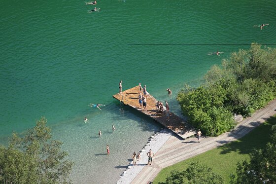 Überflugfoto Ein klarer grüner See mit Menschen, die schwimmen und auf einem Holzsteg stehen. Umgeben von Bäumen und einer schönen Landschaft.