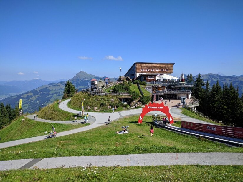 Berghütte mit Aussichtsterrasse, befestigten Wanderwegen und einem roten Torbogen auf einer grünen Alm.