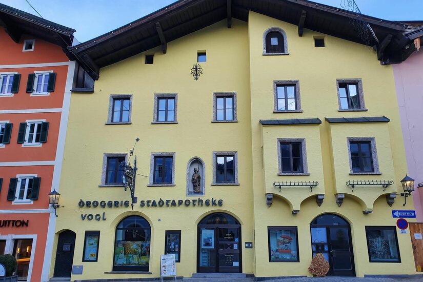Gelbe Fassade der Drogerie Stadtapotheke Vogl mit Erkern und Rundbogentüren in einer gepflasterten Straße.