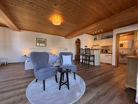 Modernes Studio-Apartment mit Holzbalkendecke, zwei blauen Sesseln, Küchenzeile und Doppelbett.